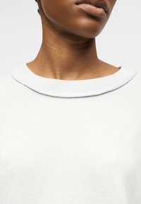 Zbliżenie osoby ubranej w jasnoszary sweter z okrągłym dekoltem, pokazujące szyję, dolną część twarzy i górną część klatki piersiowej.