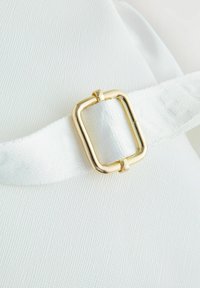 Tissu blanc avec une surface texturée, ornée d'une boucle rectangulaire dorée sur une sangle blanche. La sangle présente un motif de marque subtil.
