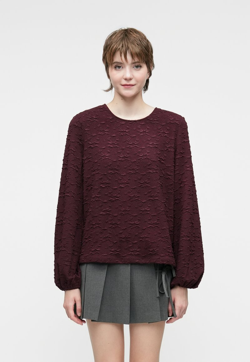 Top a maniche lunghe color marrone bordeaux con motivo floreale testurizzato, collo rotondo e vestibilità comoda. Abbinato a una gonna grigia plissettata con fiocco laterale.