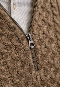 Brauner gestrickter Pullover mit strukturiertem Design, mit einem metallischen Reißverschluss an der Vorderseite und einem hell gestreiften Hemd darunter.