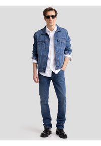 Férfi fekete napszemüvegben, kék farmerdzsekiben és farmernadrágban, fehér ingben, fekete bakancsban, egyik kezét a zsebében tartva áll.