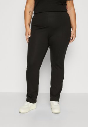 Osoba o większych rozmiarach ubrana w czarne dopasowane spodnie i białe sneakersy na platformie, stojąca na jednolitym tle.