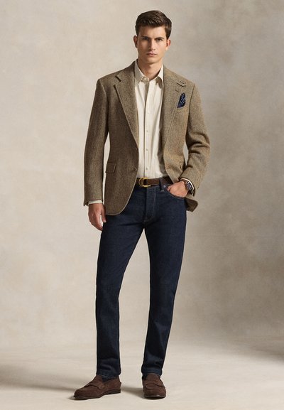 Rudas herringbone stiliaus švarkas, dryžuota balta ir smėlio spalvos marškinėliai, tamsios džinsinės kelnės, rudi zomšiniai loaferiai, mėlynas kišenėlės skarelės akcentas, stovinti poza.