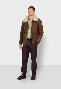 Giacca bomber in suede marrone con colletto in shearling beige, tasche frontali e dettagli neri, indossata su una camicia verde chiaro e pantaloni scuri.