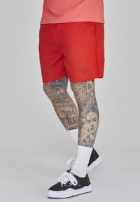 Raudoni šortai, pagaminti iš lengvos medžiagos, su logotipu ant krašto. Dėvimi su juodais sportiniais bateliais ir baltais kojiniais, demonstruojantys sudėtingus kojų tatuiruotes.