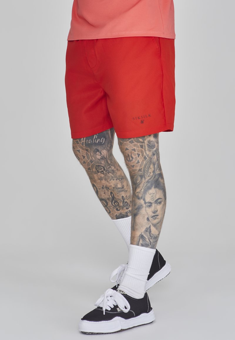 Raudoni šortai, pagaminti iš lengvos medžiagos, su logotipu ant krašto. Dėvimi su juodais sportiniais bateliais ir baltais kojiniais, demonstruojantys sudėtingus kojų tatuiruotes.