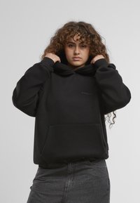 Schwarzer Hoodie mit großer Fronttasche, verstellbarem Kordelzug an der Kapuze und gerippten Bündchen. Der Stoff hat eine glatte Textur und verfügt über subtile Logodetails.