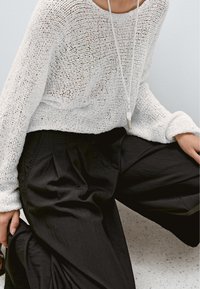 Person mit locker sitzendem weißem Strickpullover und weitem schwarzen Hosen, sitzt mit einer Hand auf dem Bein ruhend, mit sichtbarem Anhänger an einer Halskette.