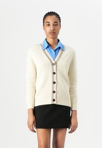 Cream knit cardigan with a textured pattern, beige trim, and dark buttons, paired with a light blue collared shirt and black skirt.