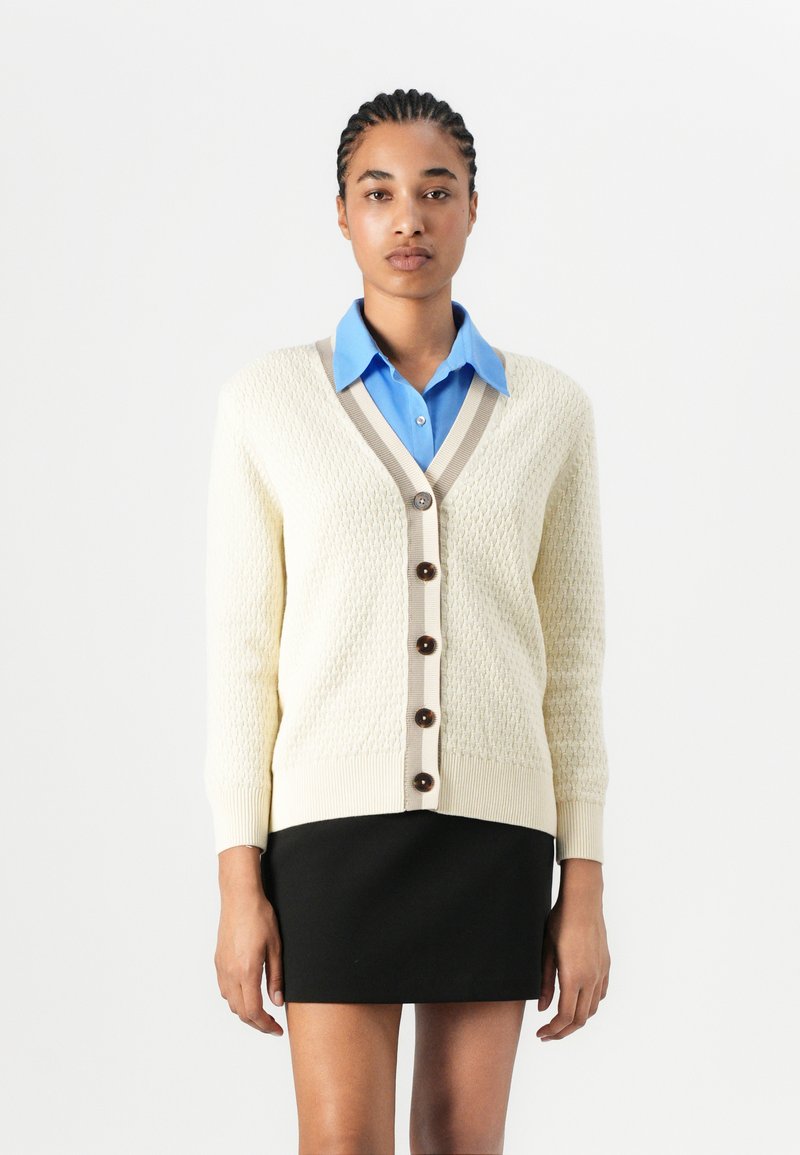 Cream knit cardigan with a textured pattern, beige trim, and dark buttons, paired with a light blue collared shirt and black skirt.