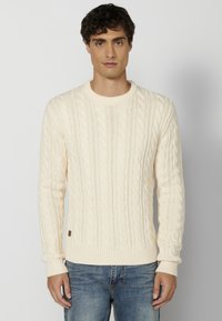 Suéter de punto trenzado en color crema con cuello redondo, puños y dobladillo acanalados, y una pequeña etiqueta marrón en el lado izquierdo, llevado sobre unos vaqueros azules.