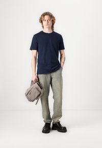 Camisa de algodón navy, jeans rectos de color verde claro, zapatos gruesos negros, sosteniendo una bolsa de lona gris con cremallera y correa ajustable.