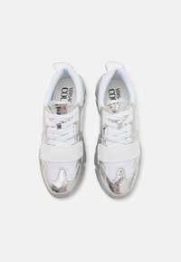 Sneakers bianchi sportivi con accenti metallici argento, texture liscia e una banda con logo prominente. Progettati con mesh e suola in gomma supportiva.