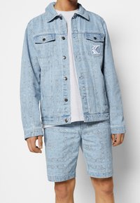 Lichtblauwe denim jack en korte broek set met een paisley patroon, knoopsluitingen en een logo-patch op de borst van het jack.