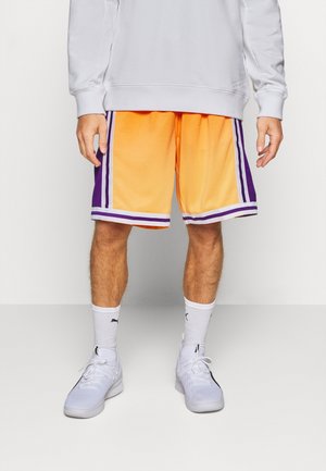 Homme portant un short de basket orange avec des rayures violettes et blanches, des chaussettes blanches, des baskets blanches et un sweat-shirt gris clair.