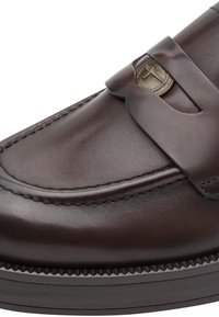 Mocassin en cuir marron foncé avec un bout rond, coutures détaillées et décoration en pièce métallique sur la bride du dessus.