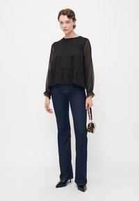Frau trägt eine durchsichtige schwarze Bluse mit langen Ärmeln, dunkelblaue Jeans, schwarze spitze Absätze und hält eine kleine Handtasche mit Leopardenmuster.