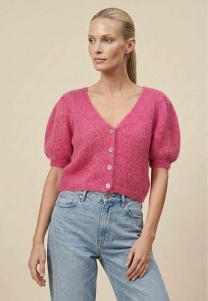 Donna con capelli biondi, maglia rosa pelosa con bottoni e maniche a sbuffo e jeans azzurri, che guarda di lato su sfondo semplice.