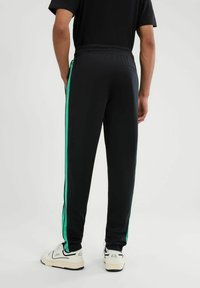 Zwarte sportieve sweatpants met groene zijkanten, elastische tailleband en een achterzak, gecombineerd met witte sneakers. Gladde textuur.