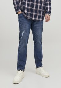 Jean en denim bleu foncé avec une déchirure usée au genou, assortis à des baskets blanches. La chemise présente un motif à carreaux bleu marine et blanc.