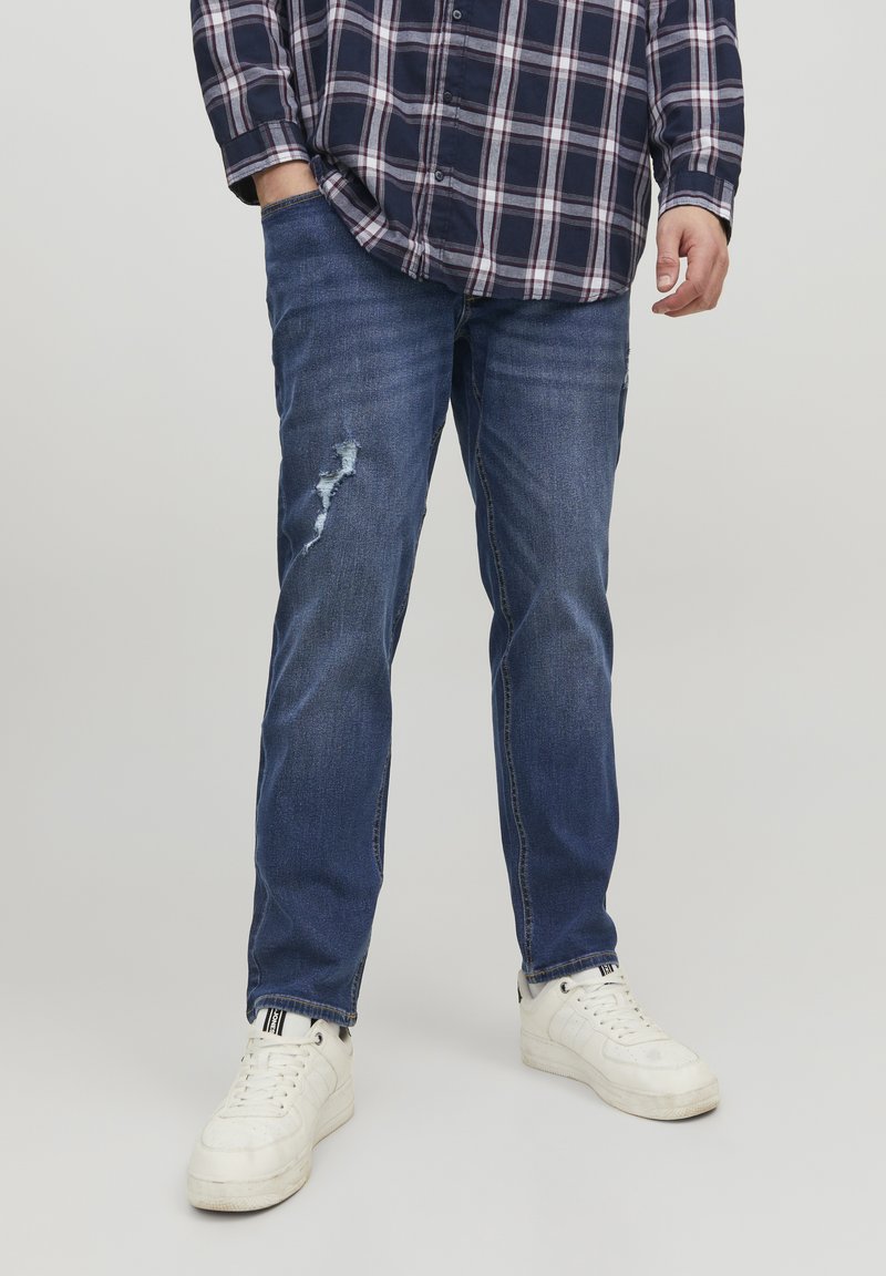 Jean en denim bleu foncé avec une déchirure usée au genou, assortis à des baskets blanches. La chemise présente un motif à carreaux bleu marine et blanc.