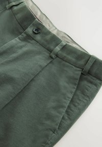 Dunkelgrüne Hose mit glatter Textur, versehen mit einem Knopfverschluss, Gürtelschlaufen und vorderen Falten. Futter im Taillenbund in heller Farbe.