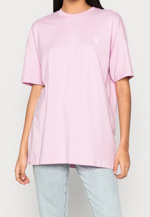 Camiseta de algodón rosa con mangas cortas, corte relajado y un pequeño logo de símbolo femenino blanco en el lado izquierdo del pecho. Textura suave y diseño casual.