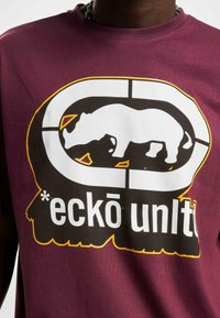 Burgunder t-skjorte laget av bomull med stort, trykket sort-hvitt neshorndesign, omgitt av en sirkulær grafikk og teksten "eckō unltd."