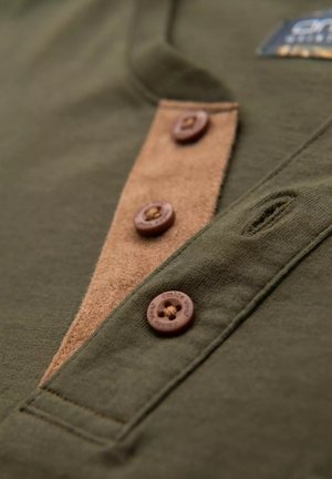 Olivgrünes Hemd mit einer braunen Wildlederblende, fünf braunen Knöpfen und dezenten Nähdetails. Weicher Baumwollstoff mit Texturvariationen.