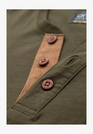 Olivgrünes Hemd mit einer braunen Wildlederblende, fünf braunen Knöpfen und dezenten Nähdetails. Weicher Baumwollstoff mit Texturvariationen.