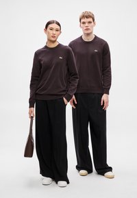 Casacos castanho-escuros de malha com gola redonda, com um pequeno logótipo verde, combinados com calças pretas de perna larga e sapatos brancos.