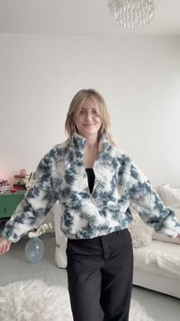 Γυναίκα που χαμογελά, φοράει μια φούτερ ζακέτα με μαύρο και λευκό μοτίβο, στέκεται σε ένα φωτεινό σαλόνι με καναπέ, πράσινη ντουλάπα και πολυέλαιο.