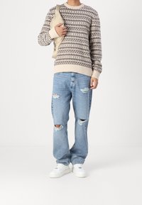 Stickad tröja i beige med bruna geometriska mönster, kombinerad med slitna ljusblå jeans och vita sneakers, hållande en beige väska.