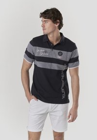 Polo rayé bleu marine et gris avec col, logo latéral et détails textuels. Associé à un short blanc ; les manches courtes sont roulées.