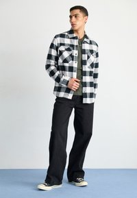 Camisa de franela a cuadros en blanco y negro, con dos bolsillos en el pecho, camiseta verde debajo, jeans de pierna ancha negros y zapatillas negras. Fondo sencillo.
