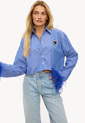 Chemise courte à rayures bleues et blanches avec un devant à boutons, ornée d'accents en plumes sur les manches et d'une broche en forme de cœur sur la poitrine.