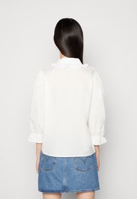 Blusa branca de mangas longas com um colarinho frisado e mangas bufantes, combinada com uma saia de ganga com costura e dois bolsos traseiros.