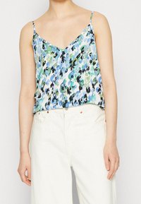 Camisola floreale in azzurro chiaro, verde e nero, con scollatura a V, spalline sottili e un tessuto leggero e fluido. Abbinata a pantaloni bianchi.