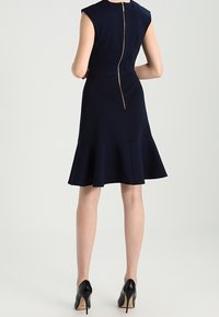 Mörkblå ärmlös klänning med en figurnära bodice, utsvängd nederkant och guldzipperdetalj bak. Bärs med svarta högklackade skor.