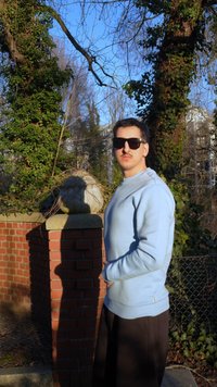 Jonge man met zonnebril en een lichtblauwe trui staat bij een stenen pilaar bedekt met klimop in een zonnige buitenomgeving.