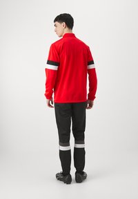 Rød idrettsjakke med svarte og hvite stripeakenter, parret med sorte bukser med lignende striper, og svarte idrettssko.
