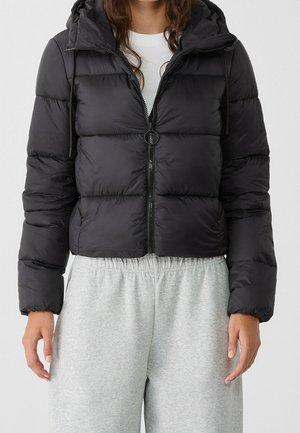 Femme portant une doudoune noire avec capuche et fermeture éclair, associée à un pantalon de survêtement gris clair et une chemise blanche en dessous.