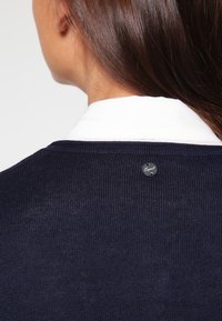 Pull en maille bleu marine avec un col blanc. Vue de dos montrant une petite étiquette noire avec logo au niveau de l'encolure. La texture semble lisse et douce.