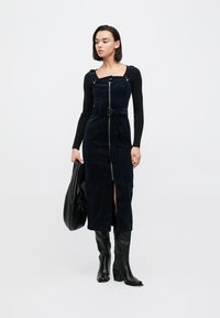 Σκούρο μπλε ρούχο από κορντούρο με φερμουάρ μπροστά και ζώνη, με ρυθμιζόμενους ιμάντες. Συνδυάζεται με μαύρο πουλόβερ με μακριά μανίκια και μπότες μέχρι το γόνατο.