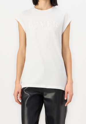 Mujer con top blanco sin mangas con el texto en relieve "LOVER PINKO" y pantalones de cuero negros, de pie frente a un fondo blanco.