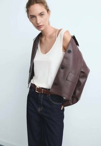 Hnědá zkrácená kožená bunda přes bílý top bez rukávů, kombinovaná s kalhotami z tmavého denimu s vysokým pasem a hnědým opaskem. Minimalistický design, hladká textura.