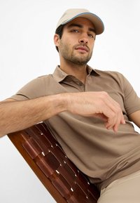 Polo beige con tessuto a trama, caratterizzato da un colletto classico. Un berretto color beige chiaro con un accento blu si trova sopra, mentre il modello si riposa in modo casual.
