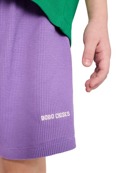 Mano di bambino accanto a pantaloncini viola con trama e la scritta "BOBO CHOSES" e una maglietta verde parzialmente visibile sopra i pantaloncini.