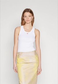 Ung jente med rett, lysebrunt hår iført en hvit ermløs topp og et gult metallisk blyantskjørt som står foran en ensfarget bakgrunn.