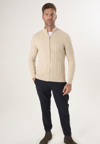 Cremfarbener, kabelgeflechter Zip-Up Cardigan, mit gerippten Bündchen und Saum, kombiniert mit marineblauen Hosen und braunen Stiefeletten. Glatte Textur, lässiges Design.